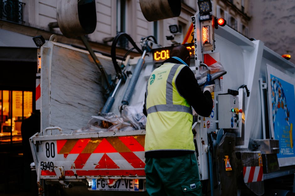 A street cleaner dressed in high-visibility orange clothing and a matching hat is sweeping a sidewalk near a wall covered with scattered litter, including paper, plastic, and food waste. The cleaner i